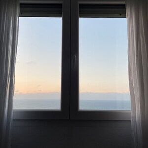 Puede incluir: Una vista del océano desde una ventana con cortinas blancas. El cielo es de un azul claro con algunas nubes. El agua es de un azul profundo y el horizonte es visible en la distancia.