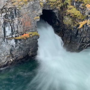 May include: A waterfall cascades through a rocky crevice, creating a misty, ethereal effect. The water is a milky white, contrasting with the dark gray rocks and the vibrant green foliage.