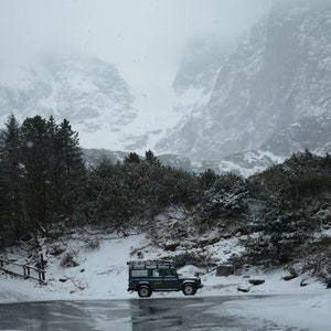 May include: A blue off-road vehicle parked in a snowy mountain landscape. The vehicle has a roof rack and is surrounded by snow-covered trees and mountains.