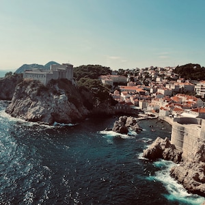 Könnte beinhalten: Blick auf eine Küstenstadt mit einer Festung auf einer Klippe, die über das Wasser ragt. Die Stadt ist an einem Hang gebaut und hat viele rote Ziegeldächer. Das Wasser ist tiefblau und im Vordergrund befinden sich Felsen.