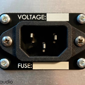 May include: Close-up of a black power outlet with a silver faceplate. The faceplate has two labels, "VOLTAGE:" and "FUSE:" with blank spaces for text. The outlet is secured with four silver screws.