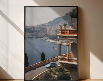 May include: Framed print depicting a coastal scene with a light blue car on a winding road. The image shows a detailed building with a balcony overlooking the water and a city in the background. The print is in a black frame.