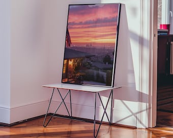 Peut inclure: Une photographie encadrée d'un coucher de soleil sur une ville. La photo est exposée sur une table blanche avec des pieds en métal noir.