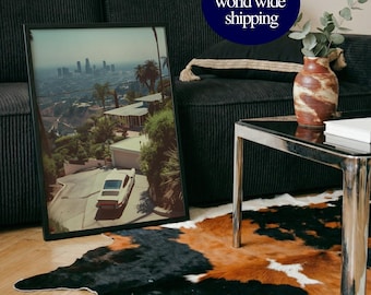 May include: Framed photograph of a vintage car driving on a road with a city skyline in the background. The photograph is in a black frame and is leaning against a black corduroy sofa. A cowhide rug with black, brown, and white patches is on the floor.