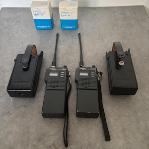 Pode incluir: Dois walkie-talkies Yaesu pretos com antenas, duas capas de couro pretas e duas caixas de bateria Yaesu FNB-22 azuis e brancas. Os walkie-talkies têm um ecrã e botões. As capas têm uma alça.
