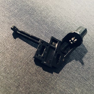May include: A black plastic component with a lever and a connector. The connector has three prongs. The component is disassembled and lying on a gray fabric surface.