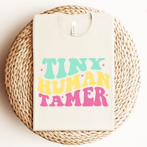 Könnte beinhalten: Ein beiges T-Shirt mit dem Text "TINY HUMAN TAMER" in einer Retro-Schriftart mit grünen, gelben und rosa Farben.