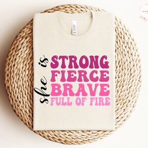May include: A beige t-shirt with the text "She is strong fierce brave full of fire" in pink and black lettering.