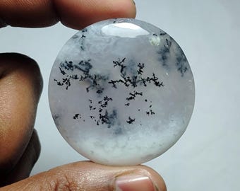 May include: A round, polished gemstone with a white and gray cloudy pattern and black dendritic inclusions. The stone is held between fingers, showcasing its smooth, reflective surface. The gemstone's design resembles a miniature landscape.