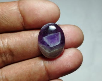 May include: An oval-shaped amethyst gemstone with a polished surface. The stone features a deep purple hue with a lighter purple and gray pattern in the center. The gemstone is held in a hand.