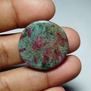 Peut inclure: Une pierre précieuse ronde avec une base turquoise et verte, parsemée d'inclusions rouges et brunes vibrantes. La pierre est tenue dans une main, mettant en valeur sa surface polie et ses motifs naturels. Cette pierre précieuse pourrait être utilisée pour la joaillerie.