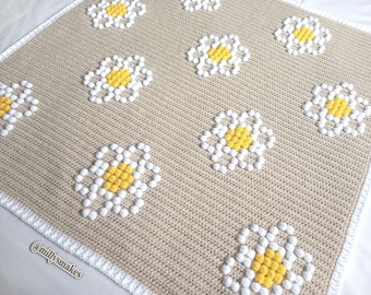 May include: A crocheted blanket with a beige background and white and yellow daisy flowers. The blanket has a white border.