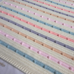 Op de afbeelding: Een wit gebreid babydekentje met strepen van roze, blauw, perzikkleurig en paars garen. Het dekentje heeft een textuurpatroon met kleine reliëf stippen.