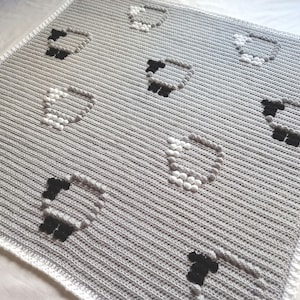 Könnte beinhalten: Eine graue Häkeldecke mit einem sich wiederholenden Muster von Schafen. Die Schafe bestehen aus weißen und grauen Knötchen für den Körper und schwarz für Kopf und Beine. Die Decke hat eine gerippte Textur und einen weißen Rand.