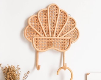 May include: A woven rattan seashell wall hook with two hooks and a matching rattan hanger with a sun design. The wall hook is mounted on a white wall and the hanger is hanging below it. The text "PLAY" is visible on a sign below the hanger.