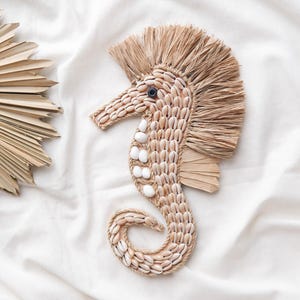 May include: A decorative seahorse wall hanging made from natural materials. The seahorse is crafted with seashells and woven fibers, featuring a textured mane and tail. The background is a white fabric with a fan-shaped decoration.
