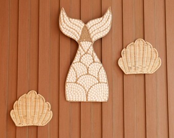 May include: A white and brown mermaid tail made of woven shells, with two smaller woven shell decorations on a brown wood background.