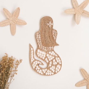Op de afbeelding: Een beige zeemeermin wanddecoratie gemaakt van geweven zeegras en witte schelpen. De zeemeermin zit met haar staart om haar heen gekruld. De decoratie is omgeven door drie beige geweven zeesterren.