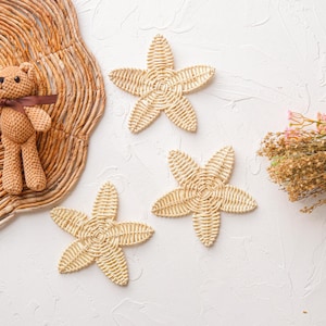 May include: Three woven star-shaped wall decor pieces in a light beige color, with a brown teddy bear plush toy on a woven mat in the background.