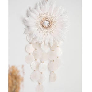 Puede incluir: Adorno de pared blanco con un diseño de flor grande y plumosa con un elemento circular central hecho de pequeñas conchas. Debajo, una cascada de discos de concha blancos y redondos cuelga de cuentas de madera.