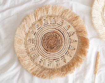 May include: A round woven placemat with a fringe border. The placemat is made of natural materials and has a white and brown pattern.