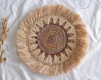 May include: A round, woven placemat with a sunburst design. The center features a spiral pattern, surrounded by a ring of rectangular shapes. The outer edge has a fringe. The colors are natural beige and brown.