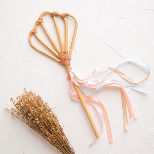 Peut inclure: Une baguette en bois avec un sommet en forme de coquille tissé et des rubans rose pâle, bleu et blanc attachés.