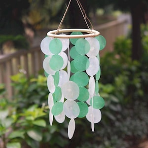 May include: A hanging wind chime with a wooden ring at the top and strands of green and white circular discs. The discs are arranged in a cylindrical shape, creating a decorative and airy design. The wind chime is suspended by a natural fiber string.