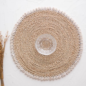 Puede incluir: Un mantel individual redondo tejido con un borde de conchas blancas. El mantel individual está hecho de fibras naturales y tiene un aspecto rústico y costero.