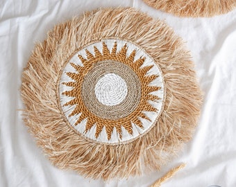 Pode incluir: Um tapete de mesa redondo tecido com uma borda de franjas. O centro do tapete de mesa apresenta um design de sol com detalhes tecidos em branco e marrom.