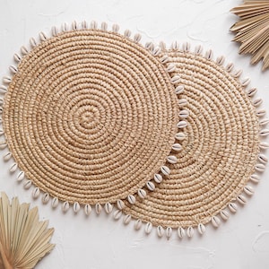 May include: Two round, woven placemats with a spiral design and a border of small, white seashells. The placemats are a natural beige color and are placed on a white surface. Dried palm leaves are also visible.