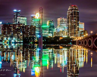 May include: Nighttime cityscape with illuminated skyscrapers and a bridge reflected in the water. The buildings feature various architectural styles and are lit with warm and cool-toned lights. The image has a long exposure, creating a mirror-like reflection.