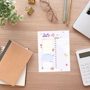 Könnte beinhalten: Ein tägliches Planungsblatt mit den Worten "Daily planner" oben, mit Abschnitten für eine To-Do-Liste, einen Zeitplan und Notizen. Das Blatt ist mit farbenfrohen Illustrationen von Erdbeeren, Blumen und anderen niedlichen Designs verziert. Ein Notizbuch, ein Laptop und ein Taschenrechner befinden sich ebenfalls auf der Holzoberfläche.