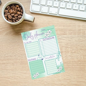 Könnte beinhalten: Ein Daily Planner Notizblock mit mintgrünem Aquarelldesign, floralen Akzenten und den Worten "Daily Planner", "To do list", "Schedule" und "Notes". Eine Tasse Kaffeebohnen und eine Tastatur sind ebenfalls zu sehen.
