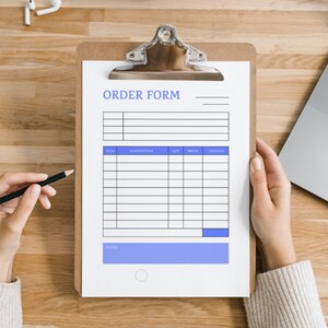 Könnte beinhalten: Ein Klemmbrett mit einem "ORDER FORM" auf einem weißen Blatt Papier. Das Formular hat Abschnitte für Artikel, Beschreibung, Menge, Preis und Betrag. Eine Hand hält einen Bleistift, bereit zum Schreiben. Ein Laptop und kabellose Ohrhörer sind ebenfalls sichtbar.
