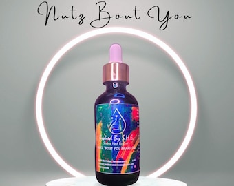 May include: A glass bottle of beard oil with a pink dropper and a colourful label that says "Badied By SHE Sisters Have Endfied 2 'BOUT YOU BEARD OIL". The bottle is sitting on a light blue platform with the words "Beard Oil" written in black cursive.