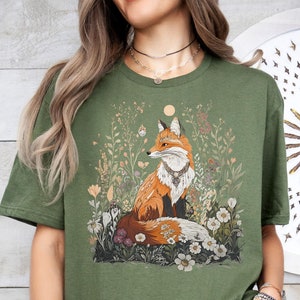 Könnte beinhalten: Ein grünes T-Shirt mit einem Aufdruck eines Fuchses, der in einem Blumenfeld sitzt. Der Fuchs ist orange und weiß mit einer schwarzen Nase und schwarzen Augen.