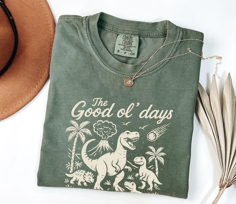 May include: Olive green t-shirt with a dinosaur graphic and the text "The Good ol' days". The shirt has a crew neck and short sleeves. A gold necklace with a pendant is draped across the shirt.