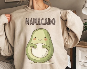 Könnte beinhalten: Ein beiger Pullover mit einem Cartoon-Avocado, der einen Kern hält, und dem aufgedruckten Text "Mamacado" auf der Vorderseite.