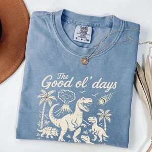 May include: A light blue t-shirt with the text "The Good ol' days" and a graphic of dinosaurs, palm trees, and a volcano. A gold necklace with a round pendant is on the shirt.