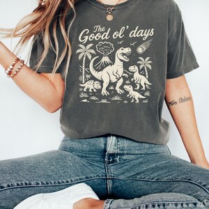 May include: Dark gray t-shirt with a dinosaur graphic and the text "The Good ol' days". The design features a volcano, palm trees, and dinosaurs. The shirt is paired with blue jeans and white shoes.