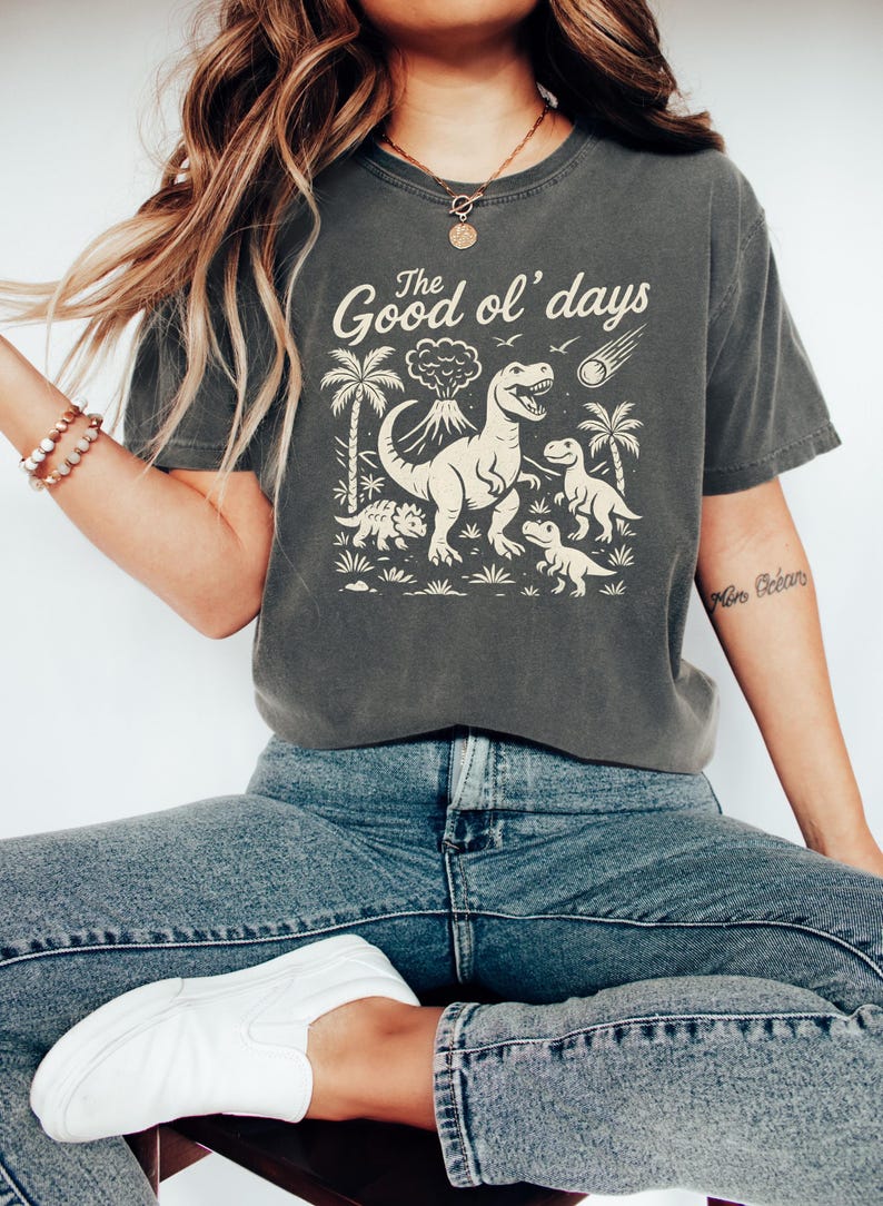 May include: Dark gray t-shirt with a dinosaur graphic and the text "The Good ol' days". The design features a volcano, palm trees, and dinosaurs. The shirt is paired with blue jeans and white shoes.