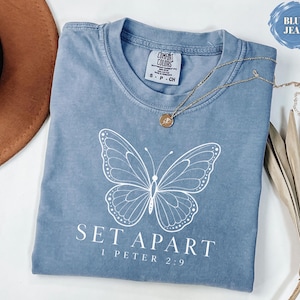 Könnte beinhalten: Ein blaues T-Shirt in Jeansoptik mit einer weißen Schmetterlingsgrafik in Linienführung auf der Brust. Unter dem Schmetterling steht der Text SET APART und 1 PETER 2:9 in weißen Buchstaben. Das T-Shirt hat einen Rundhalsausschnitt und ein Etikett.