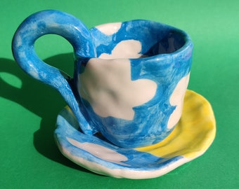 May include: A blue and white ceramic teacup with a yellow saucer. The cup and saucer are decorated with a cloud pattern.