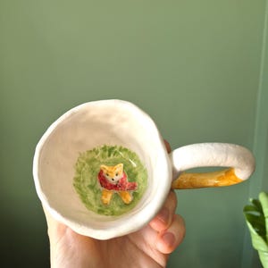 May include: White ceramic mug with a yellow handle. The inside of the mug features a hand-painted scene of a fox wearing a red scarf in a green grassy area.