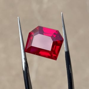 Könnte beinhalten: Ein tief roter Edelstein mit rechteckiger Form, gehalten von einer Pinzette.