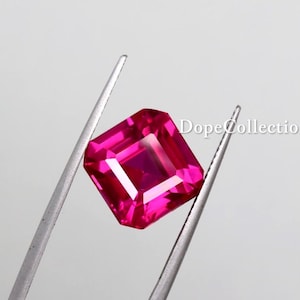 Puede incluir: Una vibrante gema rosa intenso, de corte cuadrado, es sostenida por unas pinzas plateadas sobre un fondo blanco. La gema tiene una superficie facetada, que refleja la luz. El texto "DopeCollectionIN" es visible en el fondo.