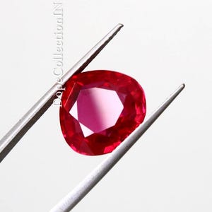 以下が含まれることがあります： 銀色の金属製ピンセットで保持された、鮮やかな赤色のファセットカットされた洋ナシ型のルビーの宝石。宝石は深い深紅の色合いと内部の光の反射を示しています。DopeCollectionINというテキストが石の横に垂直に印刷されています。