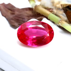 Peut inclure: Une pierre précieuse rose vif, taillée en forme ovale. La gemme est posée sur un fond blanc, avec un arrière-plan flou de feuilles et de tiges séchées. La couleur de la gemme est un rose vif et saturé, avec un haut degré de clarté.