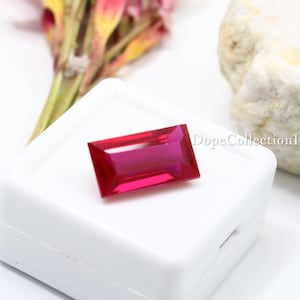 Può includere: Un rubino rettangolare di colore rosso intenso, su sfondo bianco. La gemma è sfaccettata per riflettere la luce, mostrando la sua chiarezza. L'immagine include anche il testo "DopeCollectionIN".